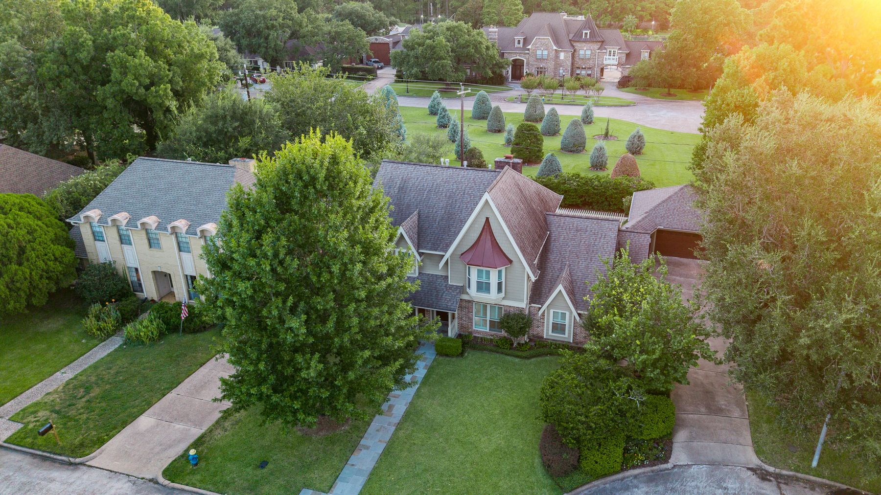 Golden-hour aerial of a two-story brick-and-siding home with a turret roof, mature trees, and a neighboring estate beyond the lawn