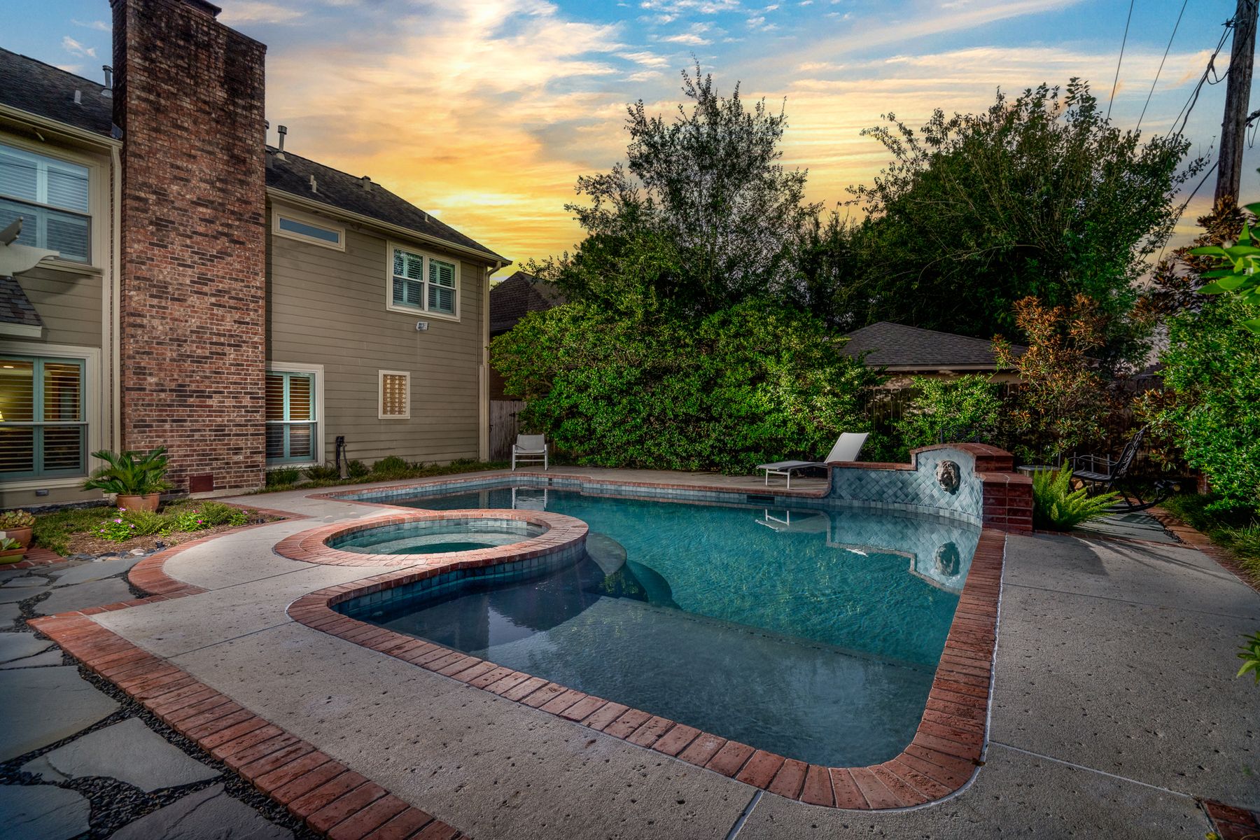 Twilight backyard with a brick-coping pool, raised spa, and a warm sunset sky behind the trees