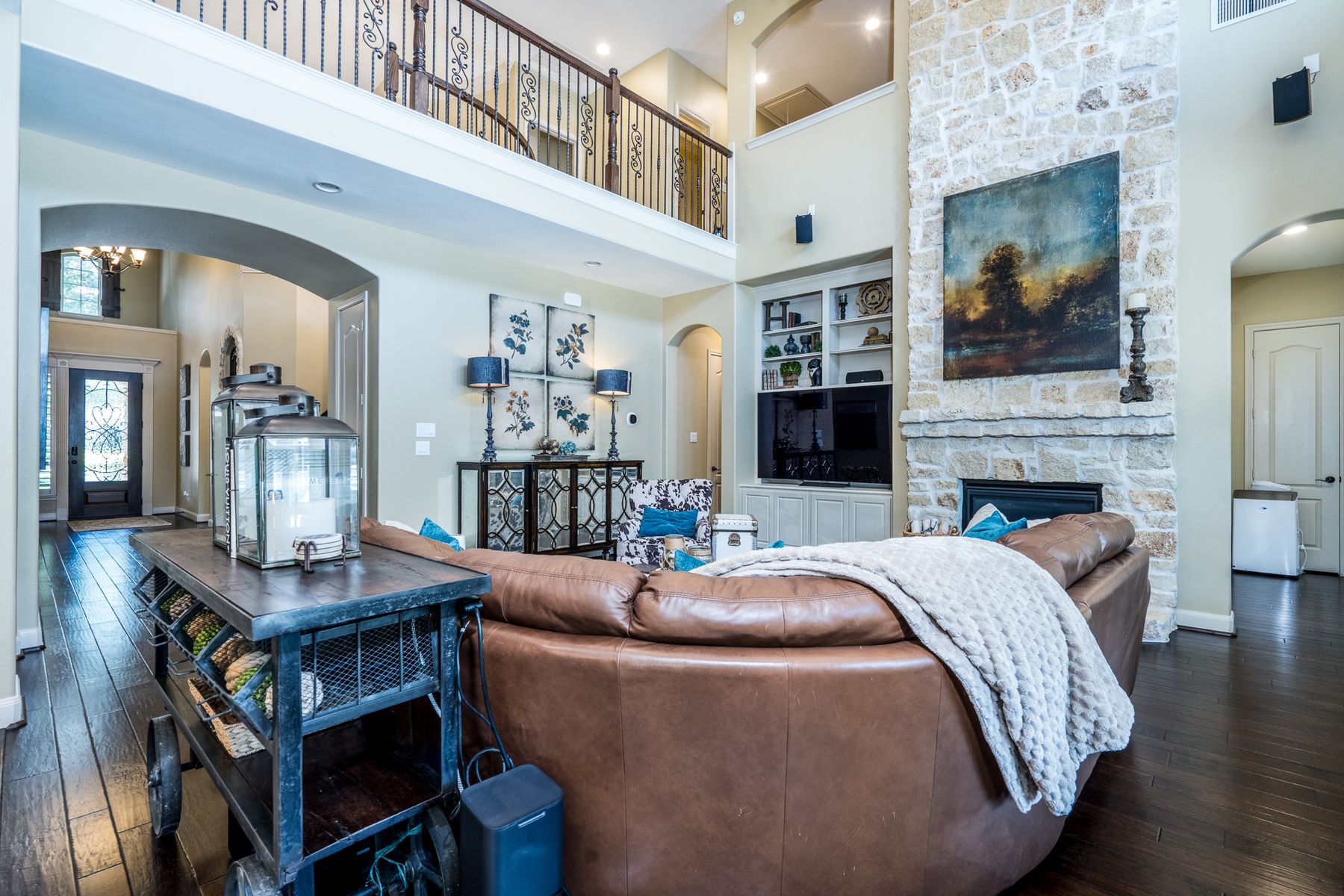 View from behind a leather sofa toward a limestone fireplace, wrought-iron balcony, and the front entry