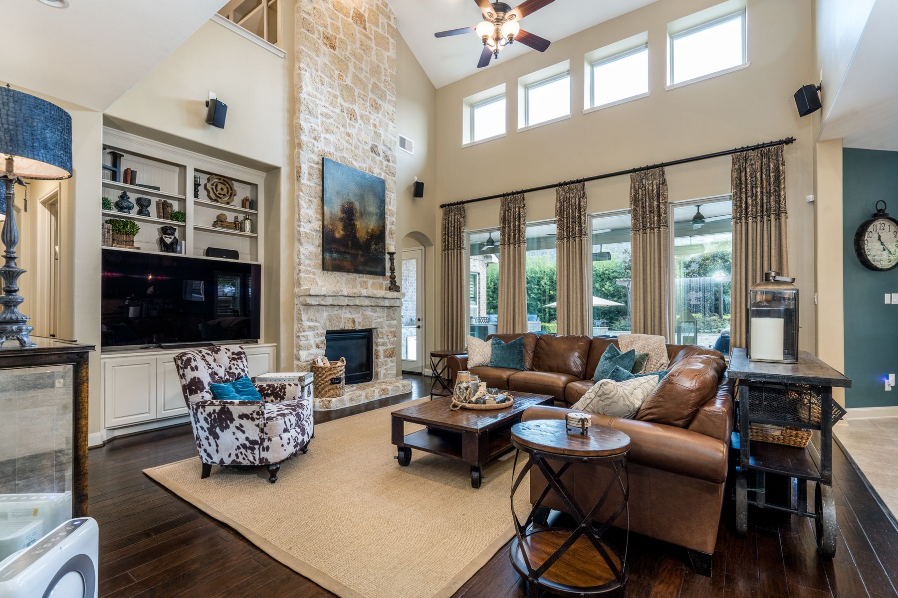 Two-story living room with a limestone fireplace, leather sectional, cowhide chair, and tall windows to the backyard