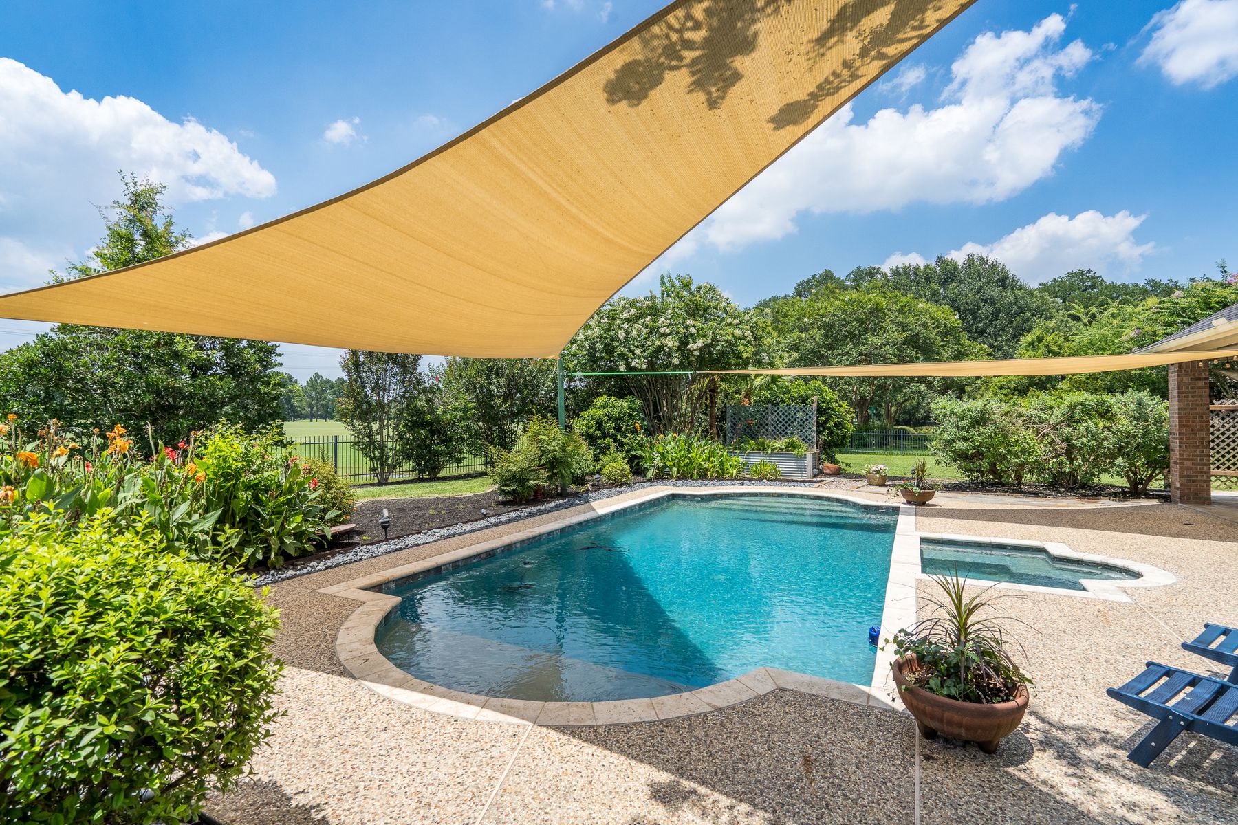 Freeform pool and spa under a tan shade sail, with a golf-course view beyond a black iron fence