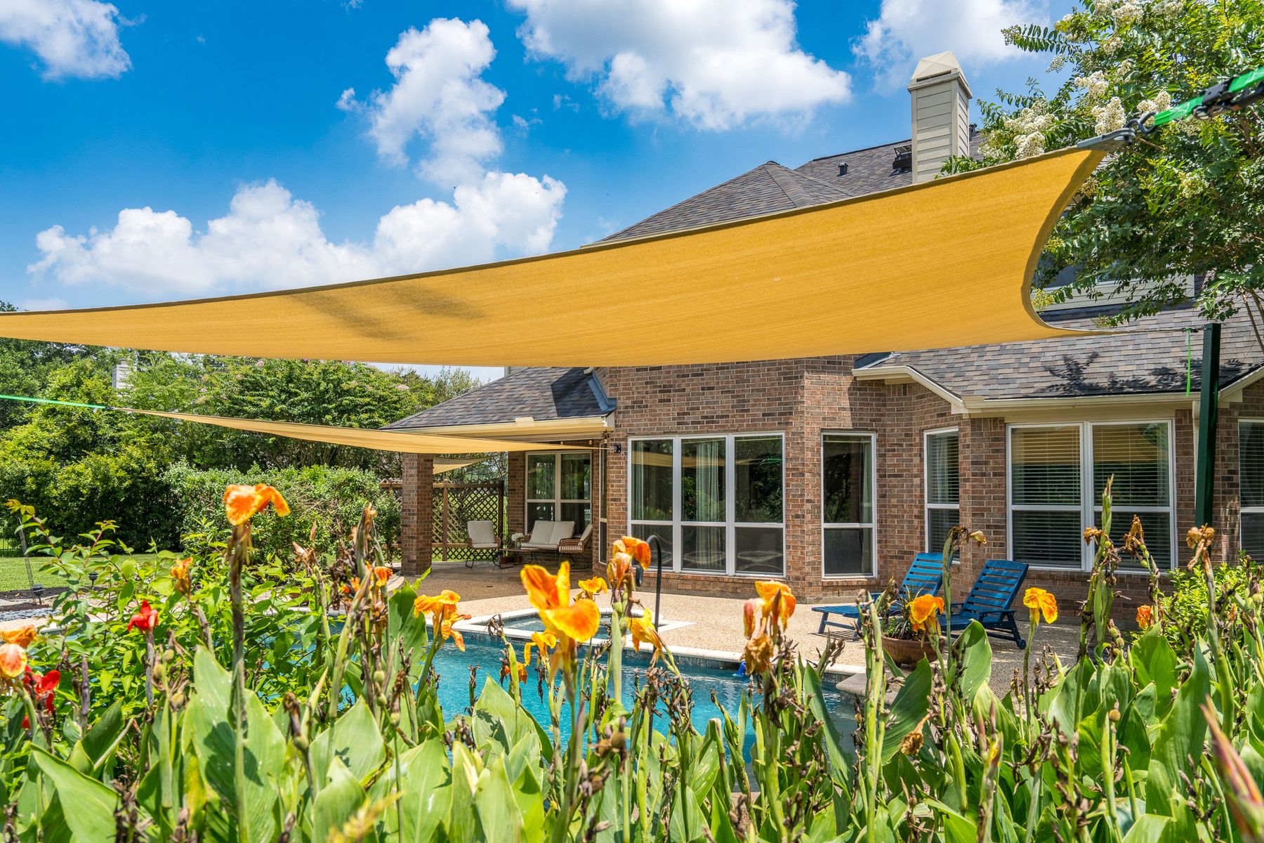 Backyard pool seen through blooming canna lilies, with a large tan shade sail stretched over a brick ranch house