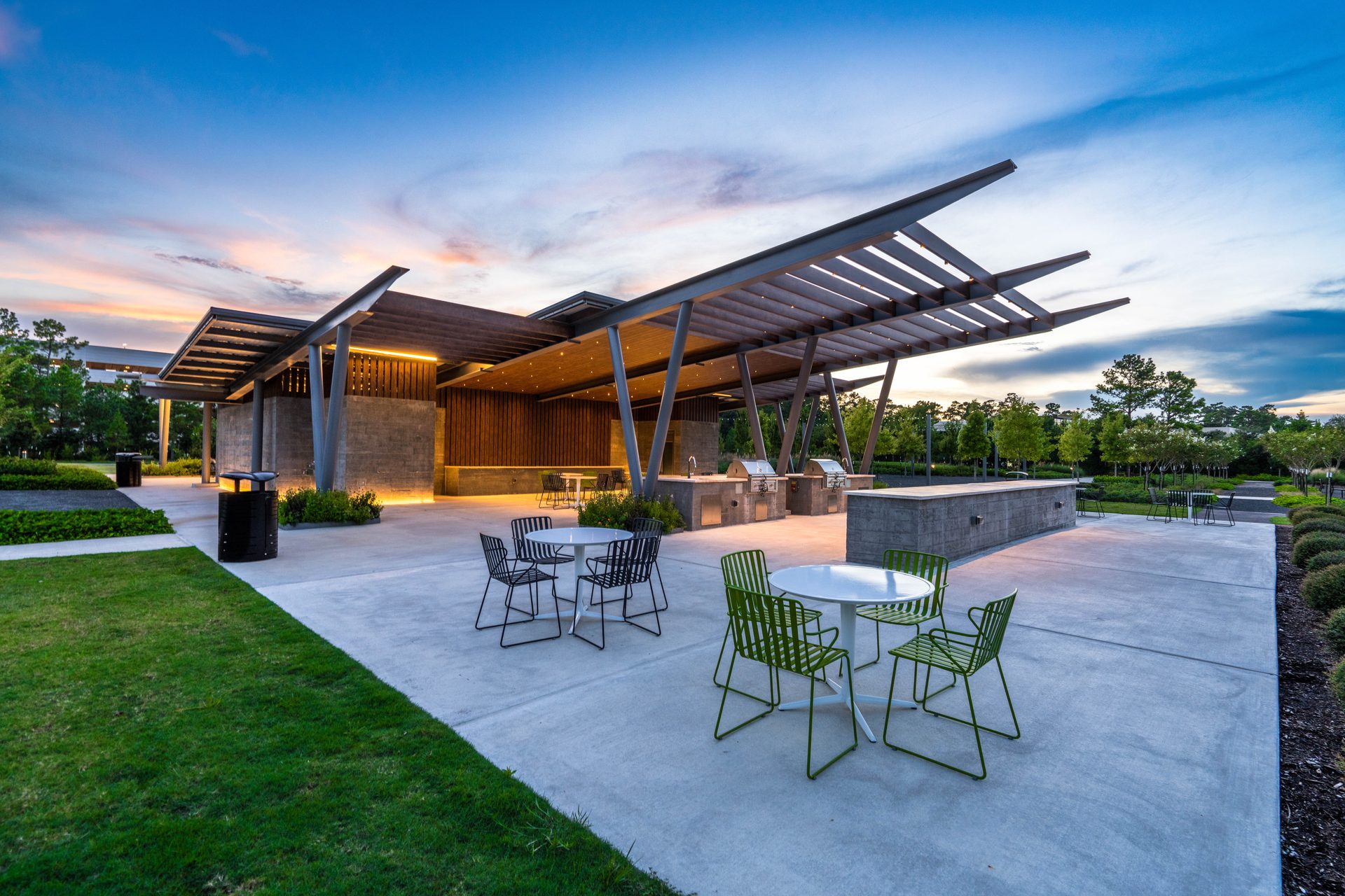 Twilight at the outdoor kitchen pavilion in Central Park, The Woodlands, with a sculptural steel pergola and lime-green cafe chairs on pale concrete